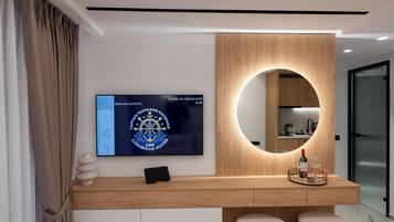A modern hotel room with a wooden console table, a large TV screen displaying a welcome message, a round mirror, and two cushioned seating areas.