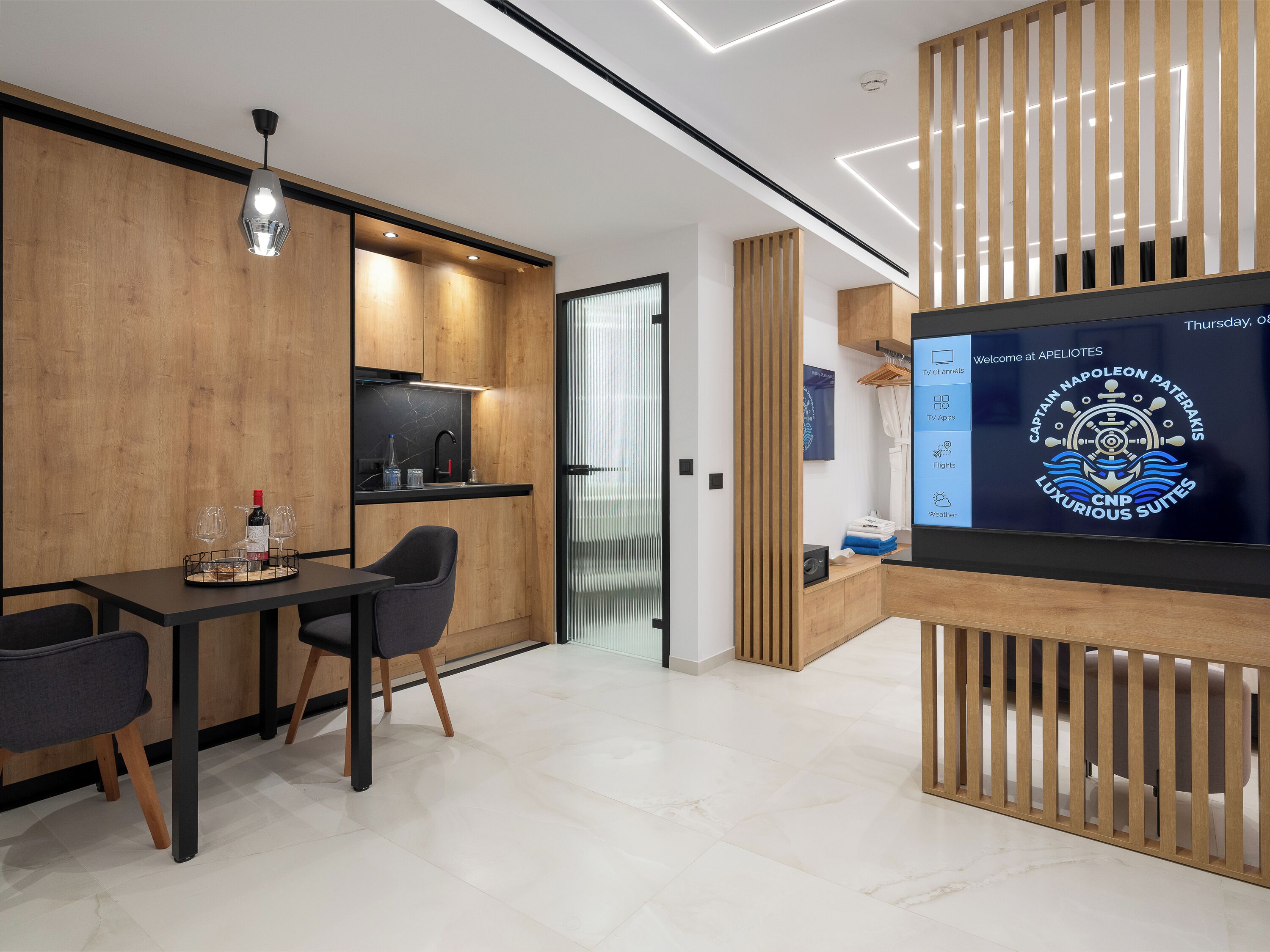 A modern hotel room with a wooden interior, a dining area with two chairs, a TV screen displaying hotel information, and a wooden partition.