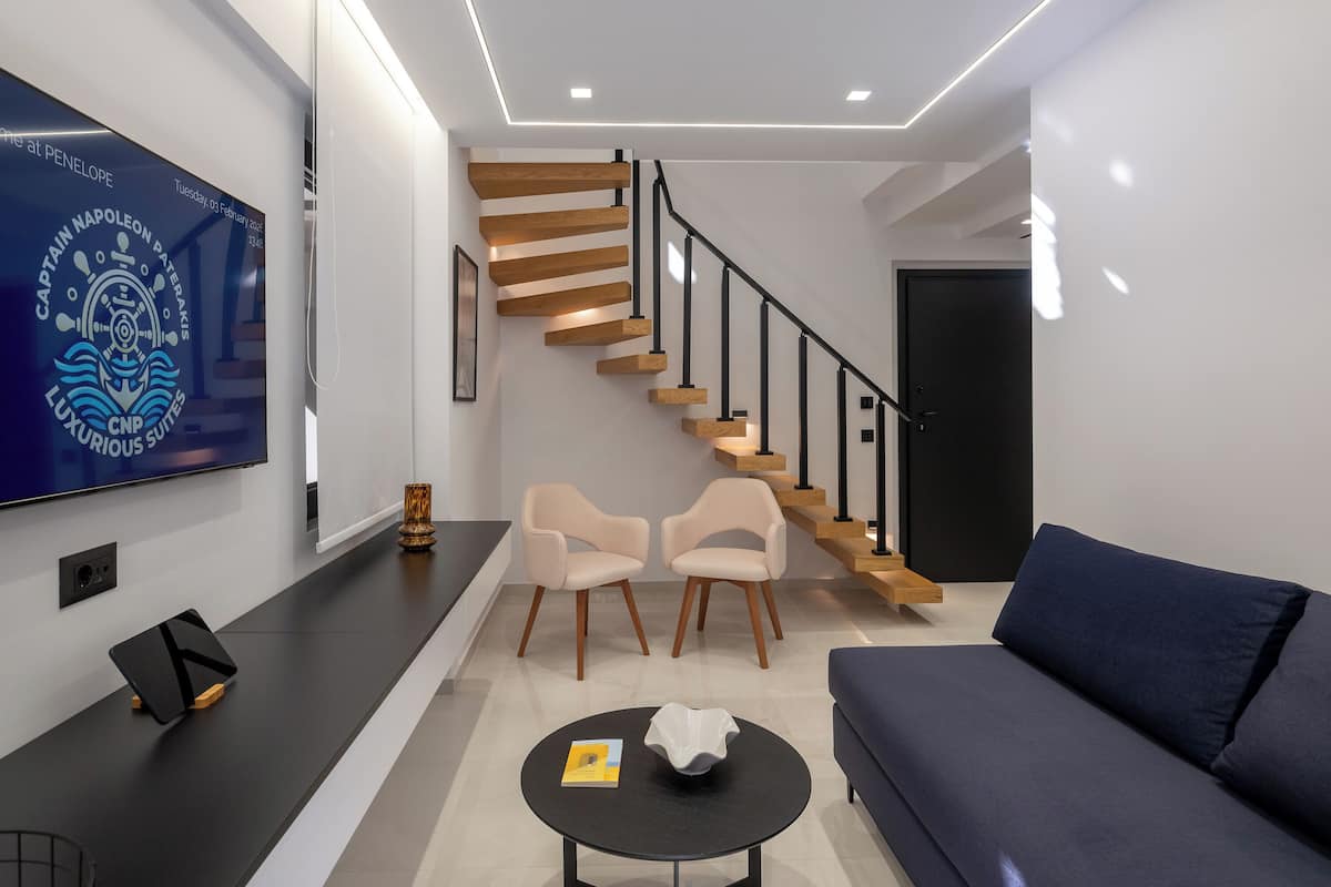 Un salon moderne doté d’un téléviseur à écran plat, d’un canapé, de deux fauteuils, d’une table basse et d’un escalier avec des marches en bois et des rampes noires.