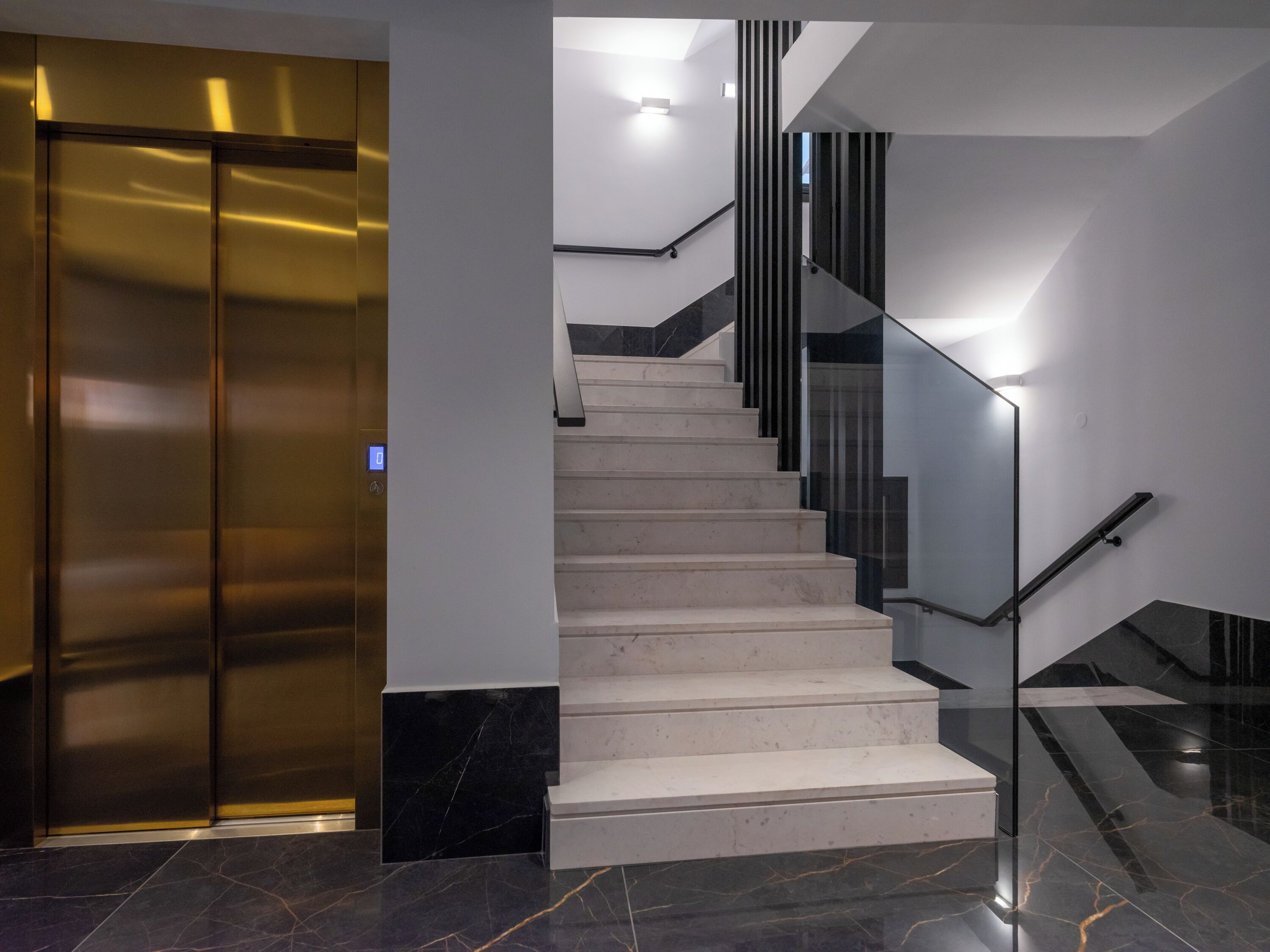 Un ascenseur moderne aux finitions métalliques, un escalier avec des rampes en verre et un revêtement de sol en marbre.