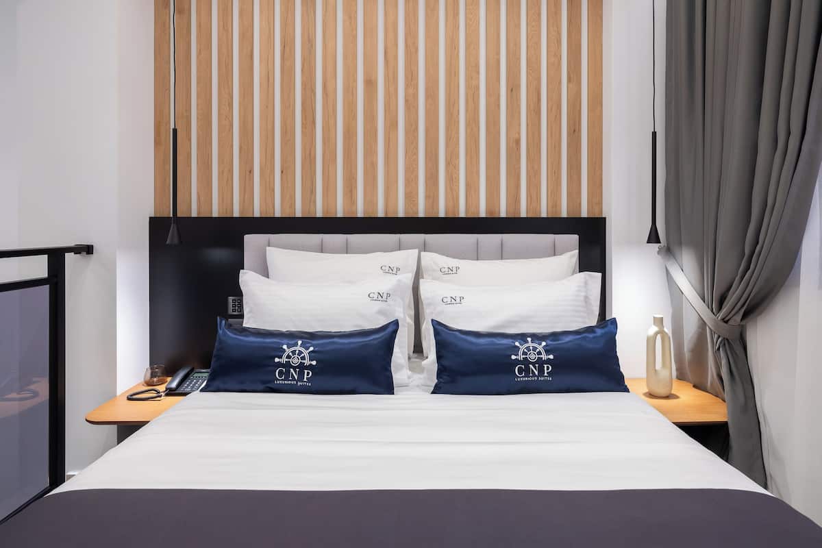 Une chambre d’hôtel avec un lit, deux oreillers portant le logo « CNP LUXURIOUS SUITES », une table de chevet avec un téléphone, une lampe et un mur avec des lambris en bois.