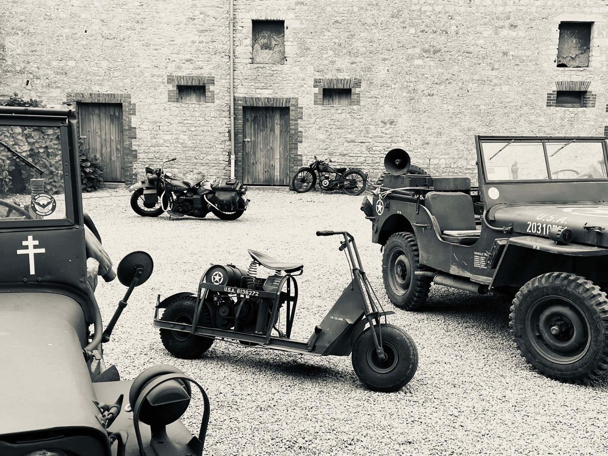 A collection of vintage military vehicles, including jeeps and motorcycles, parked in front of a stone building with multiple windows and doors.