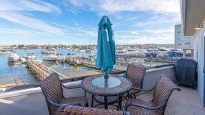 Ein Balkon mit Korbstühlen, einem Tisch und einem blauen Sonnenschirm. Blick auf einen Hafen mit vertäuten Booten.
