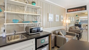 Una cocina moderna con microondas integrado, un lavamanos y un refrigerador. Hay estantes sobre la encimera con platos y vasos. Un letrero en la pared dice 'Welcome to the Beach'.