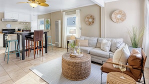A modern living room with a sofa, coffee table, and chairs. There is a kitchen area with cabinets and a dining table.