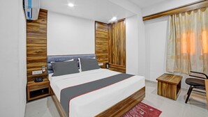 Une chambre d’hôtel moderne avec une tête de lit en bois, un lit aux couleurs sobres avec des touches de gris et de rouge, une petite table d’appoint en bois, une chaise et une fenêtre avec des rideaux.
