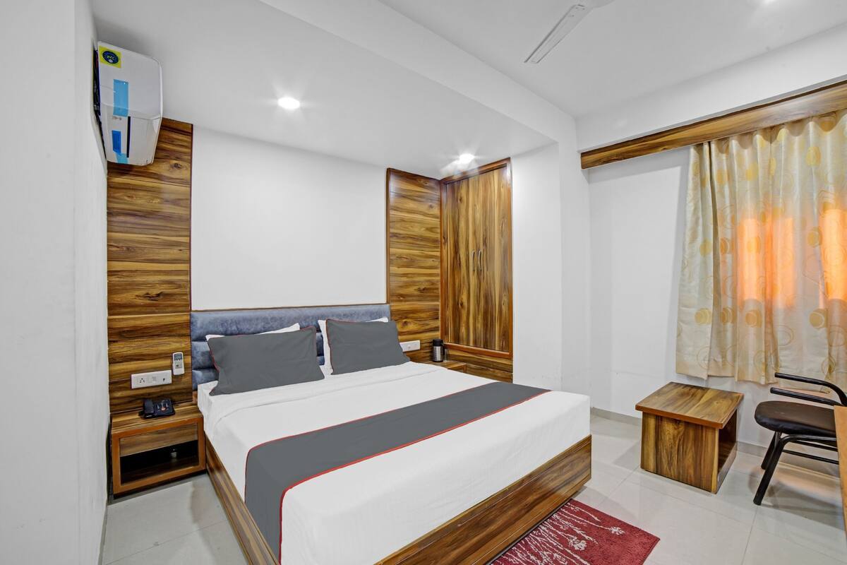 Habitació d'hotel moderna amb un panell de fusta al capçal, un llit amb detalls grisos i vermells, una taula auxiliar de fusta petita, una cadira i una finestra amb cortines.