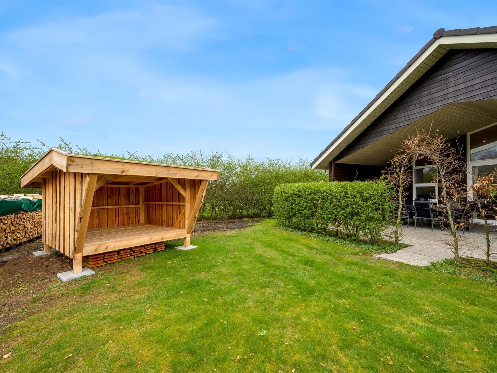 A wooden outdoor structure with a sloped roof, adjacent to a modern building with a dark roof and large windows.