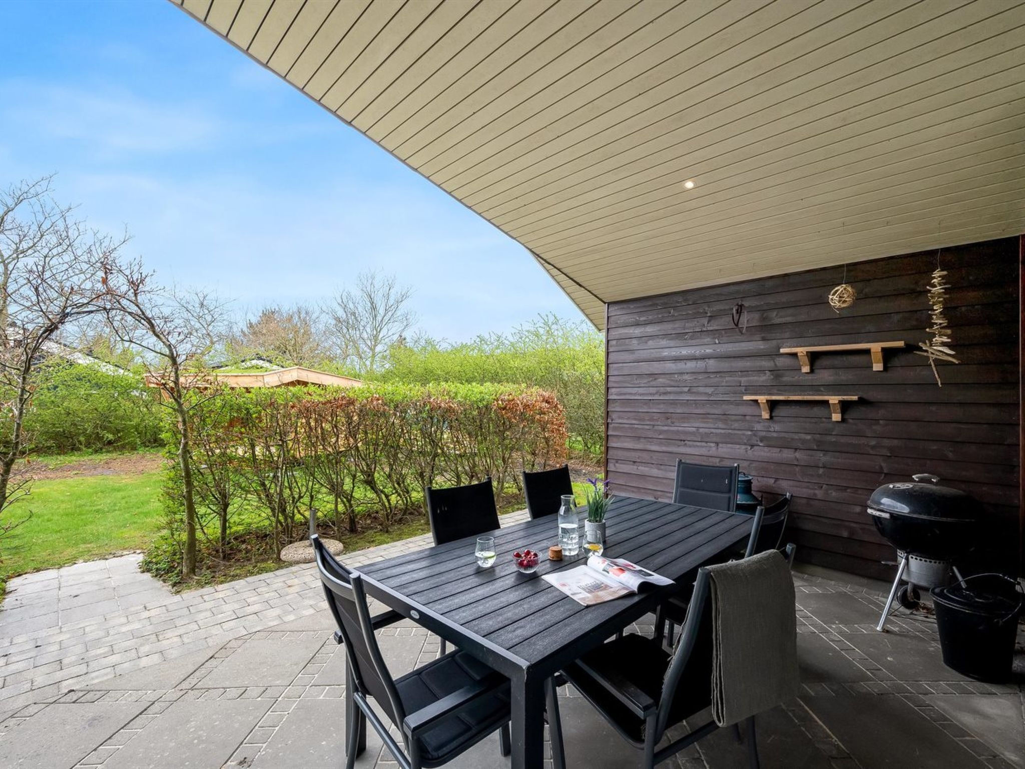An outdoor patio with a table and chairs, a barbecue grill, and a wooden wall with shelves.