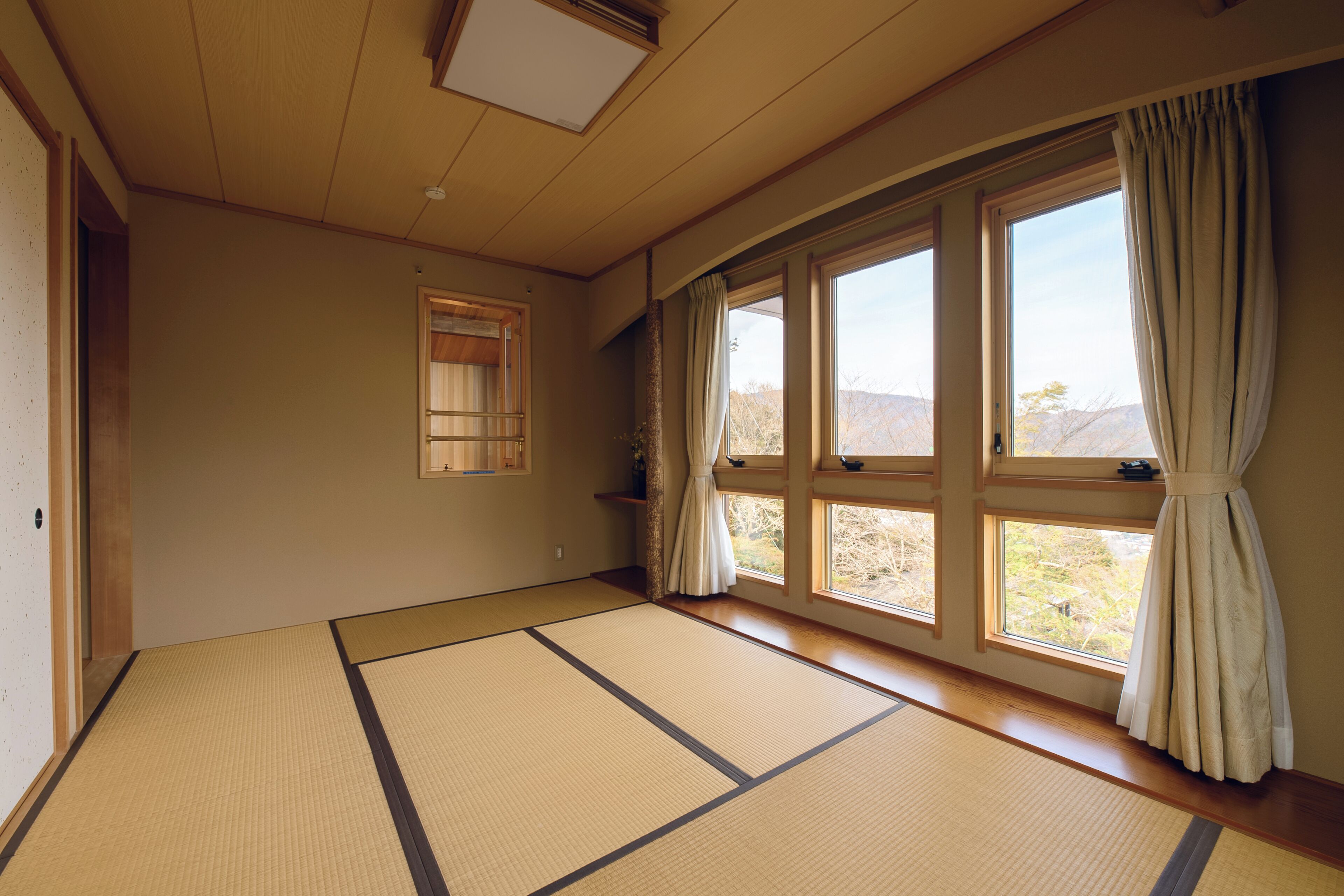 A tatami mat room with large windows, a wooden ceiling, and a sliding door.