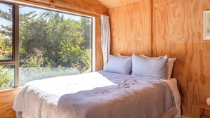 A wooden cabin bedroom with a large window, a bed with white bedding, and a wooden ceiling. - Tranquility Cottage - Central location – Walk to Beach & Village Centre (Waiheke Island)