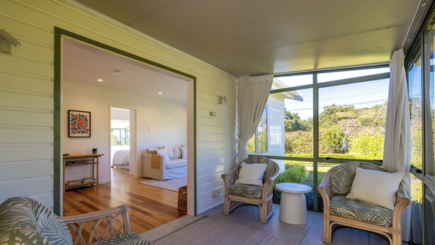 A spacious room with a large window, a wicker chair, a small table, and a sofa. - Tranquility Cottage - Central location – Walk to Beach & Village Centre (Waiheke Island)
