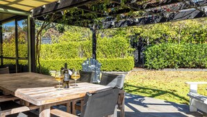 An outdoor wooden deck with a table and chairs, a grill, and a hedge in the background. - Tranquility Cottage - Central location – Walk to Beach & Village Centre (Waiheke Island)