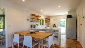 A modern kitchen with a wooden table, white chairs, and built-in shelves. - Tranquility Cottage - Central location – Walk to Beach & Village Centre (Waiheke Island)