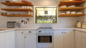 A kitchen with white cabinets, a built-in oven, wooden shelves above holding dishes, and a window with a view of greenery. - Tranquility Cottage - Central location – Walk to Beach & Village Centre (Waiheke Island)