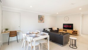 A modern living room with a dining area, a grey sofa, a TV displaying 'NETFLIX', and a small round table. - Smart Suites Self Check-in Apartments - 19 Darnley (Rocklea)