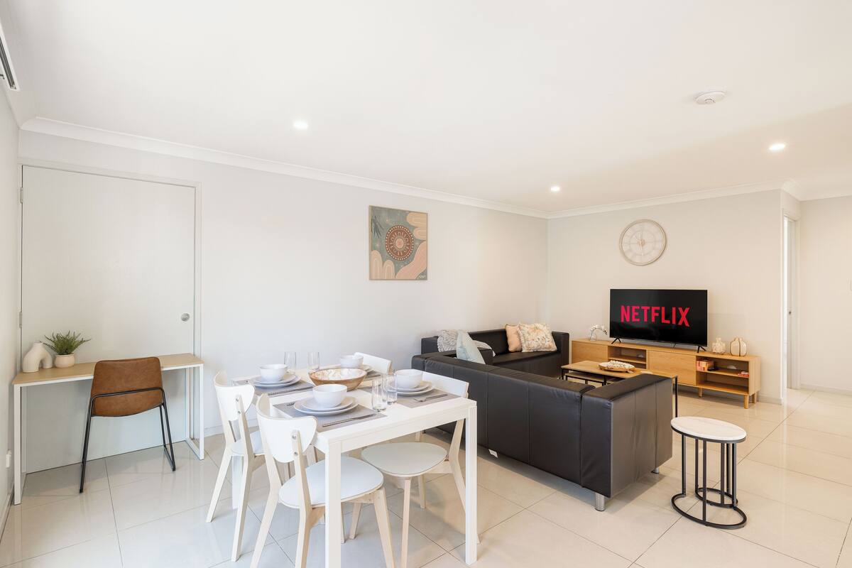 Una sala de estar moderna con comedor, un sofá gris, un televisor mostrando 'NETFLIX' y una pequeña mesa redonda.