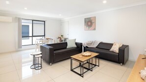 A modern living room with a black leather sofa, a wooden coffee table, and a small white table with chairs. - Smart Suites Self Check-in Apartments - 19 Darnley (Rocklea)