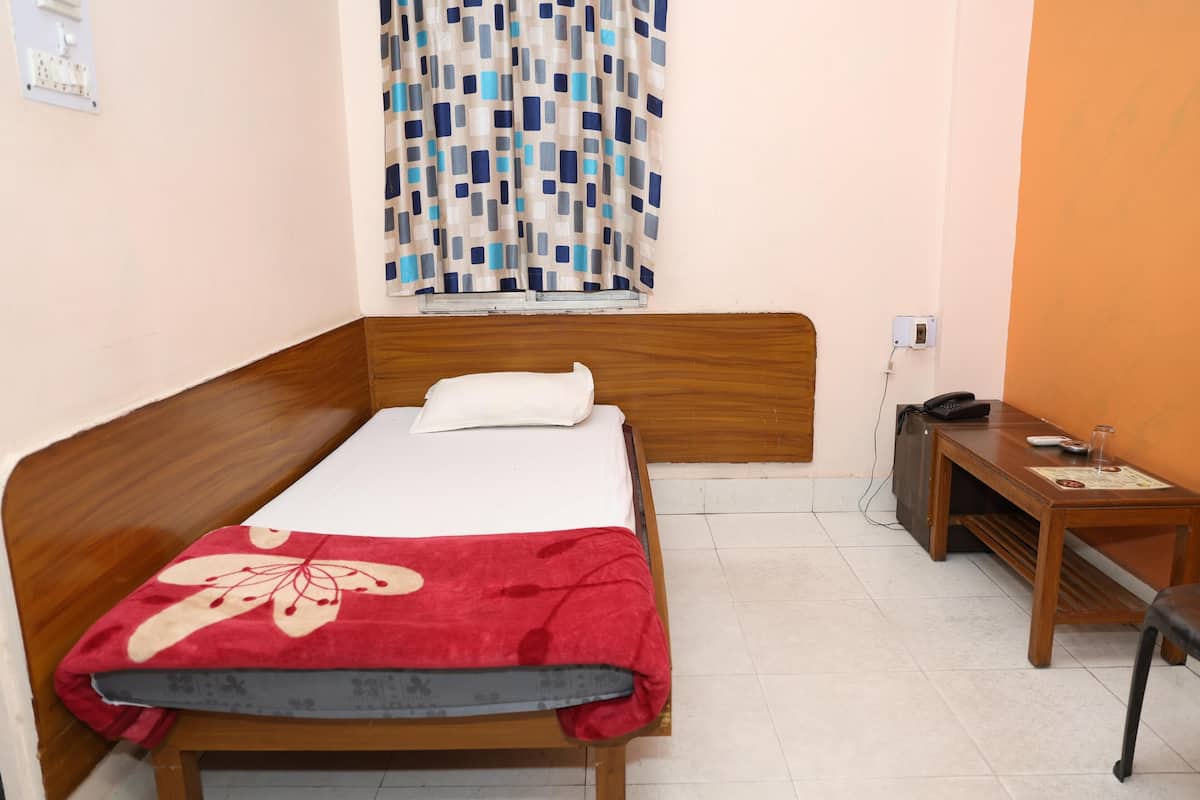 Una cama individual con cabecera de madera, una colcha roja y una almohada blanca. Junto a la cama, una pequeña mesa con un teléfono y un vaso. Una cortina estampada está parcialmente abierta en la ventana.