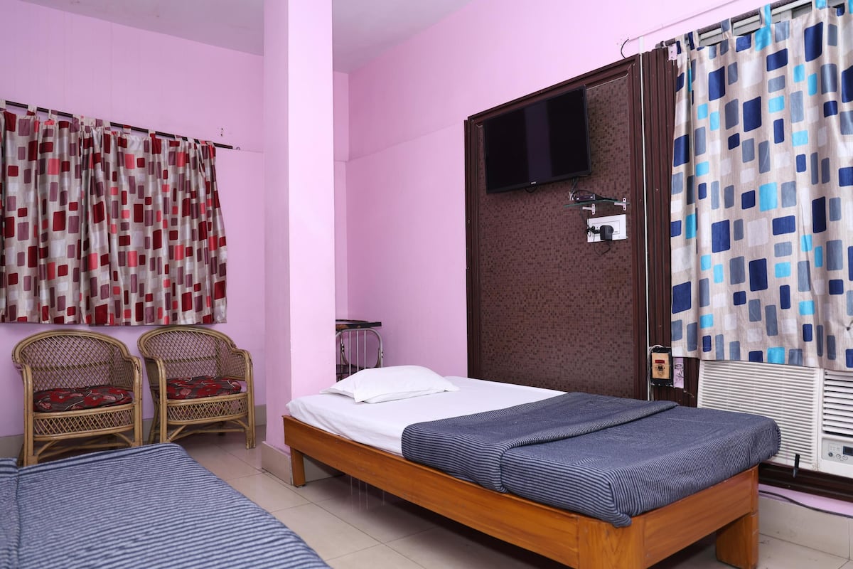 Un dormitorio con una cama individual, dos sillas de mimbre, un televisor fijado a la pared y una ventana con cortina.