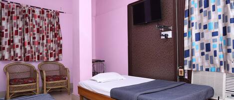 A bedroom with a single bed, two wicker chairs, a TV mounted on the wall, and a window with a curtain.