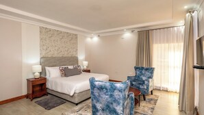 A hotel room with a bed, two armchairs, a small table, a nightstand, a lamp, a TV, and a window with curtains.