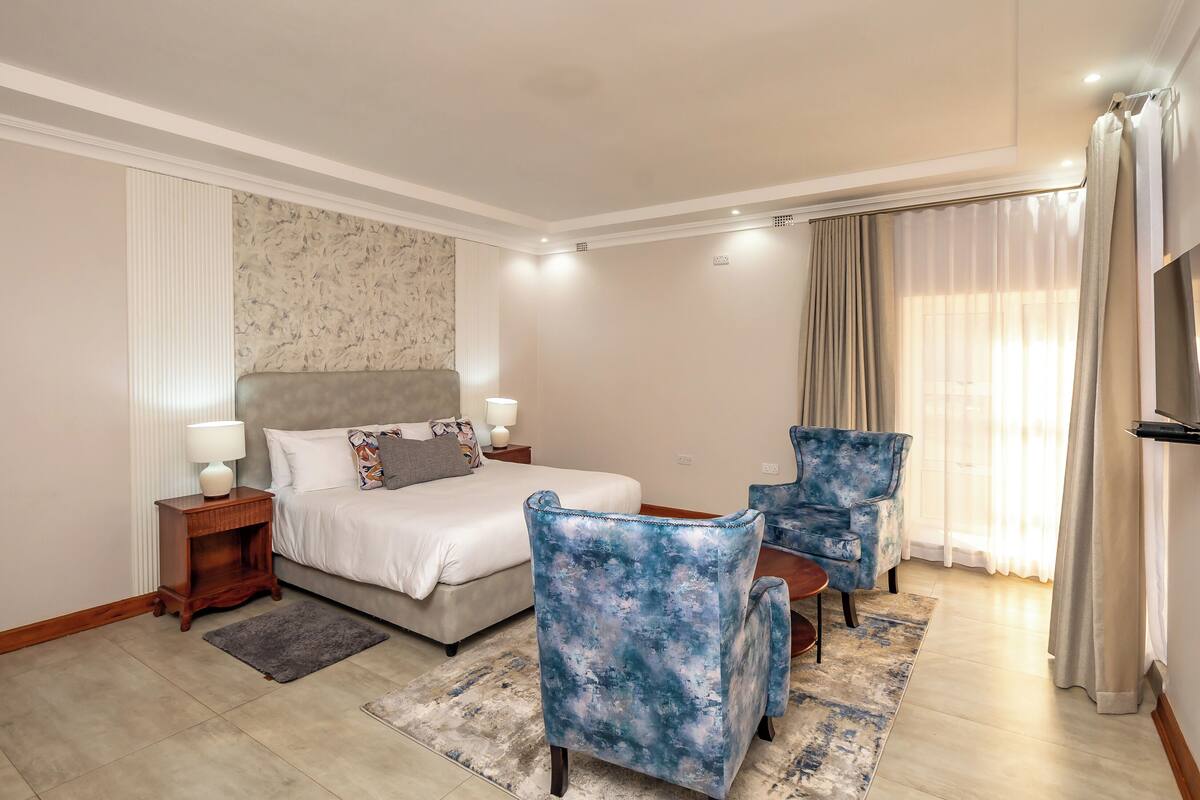A hotel room with a bed, two armchairs, a small table, a nightstand, a lamp, a TV, and a window with curtains.