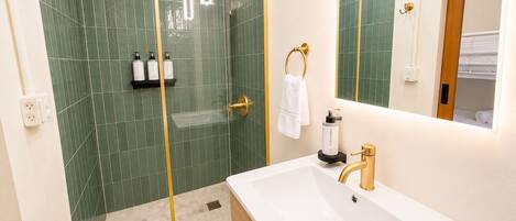 A modern bathroom with a glass-enclosed shower, a white sink with gold fixtures, and a mirror with a wooden frame.