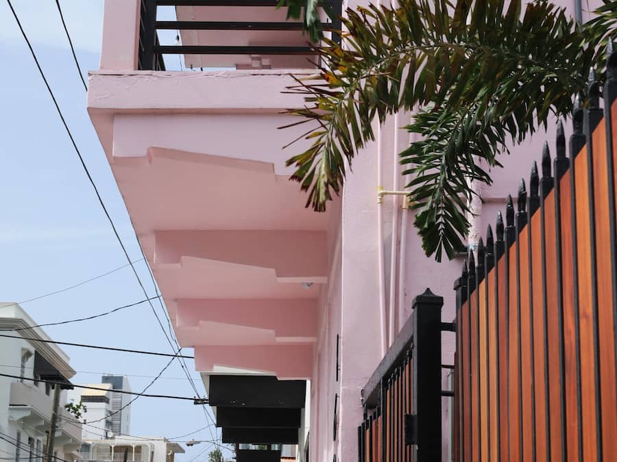 Un bâtiment rose doté d’un balcon, d’une entrée fermée par un portail et de voitures garées le long de la rue.