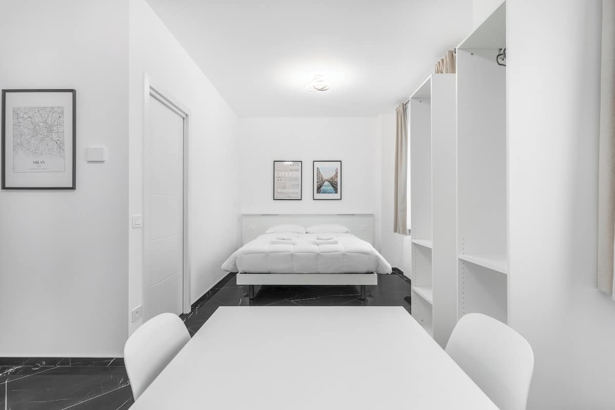 Ein modernes, minimalistisches Schlafzimmer mit einem großen Bett, einem weißen Schreibtisch und einem Stuhl. An der Wand hängen zwei gerahmte Bilder, und rechts ist ein Kleiderschrank zu sehen.