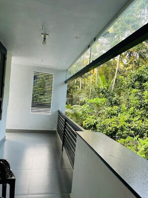A balcony with a view of dense greenery, a window with horizontal blinds, and a ceiling light fixture.