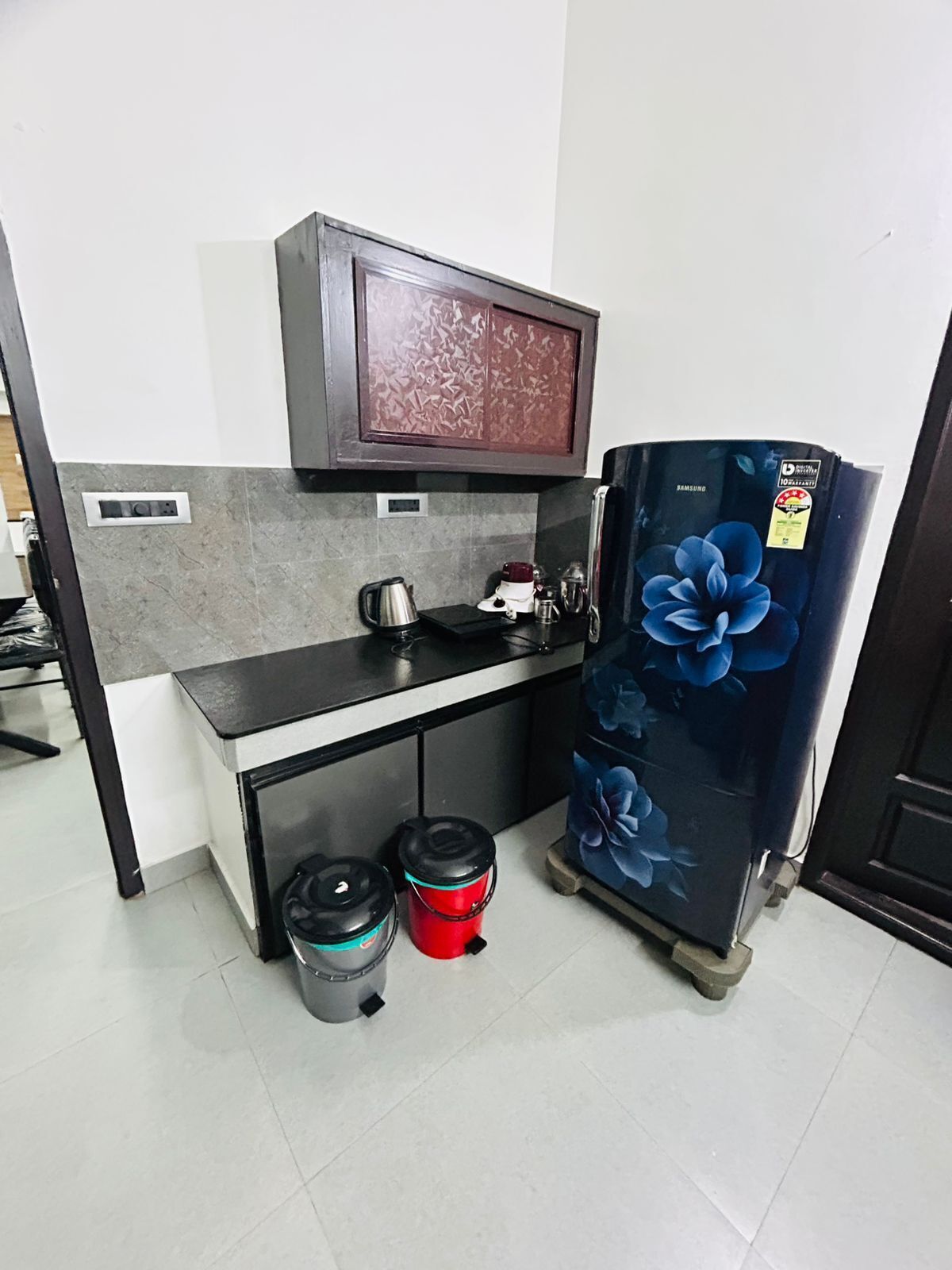 Une cuisine compacte équipée d’un micro-ondes encastré, d’un petit plan de travail, d’un réfrigérateur orné de motifs floraux et de deux poubelles.