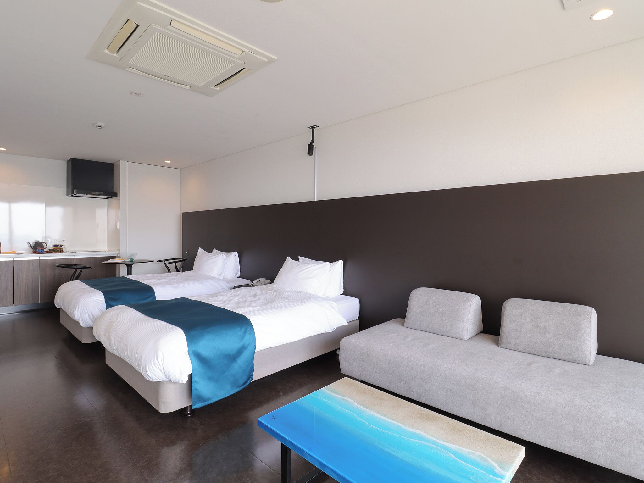 A modern hotel room with two beds, a sofa, a kitchen area, and a flat-screen TV.