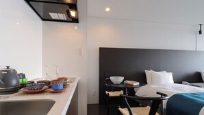 Une chambre d’hôtel moderne équipée d’une kitchenette, d’un lit avec du linge de maison blanc, d’une petite table ronde sur laquelle se trouvent un bol et un livre, et d’une chaise avec un siège en osier.