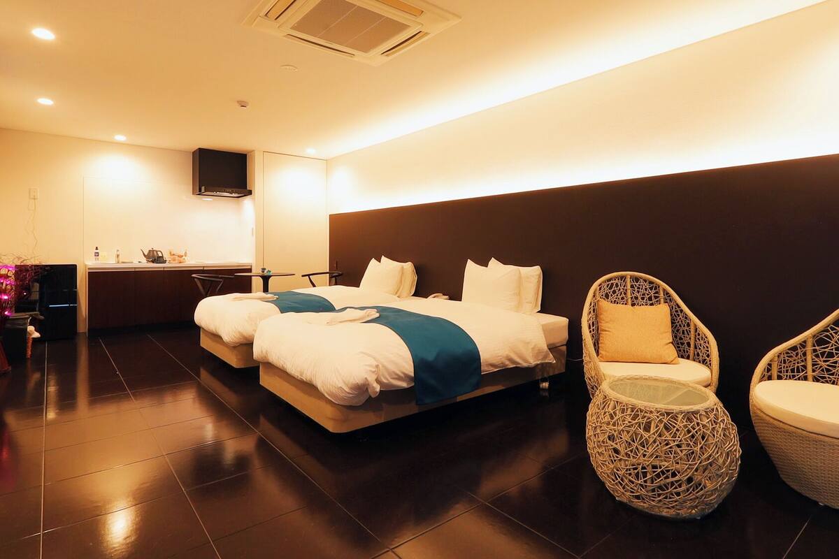 Ein modernes Hotelzimmer mit einem großen Bett, zwei Korbstühlen, einem kleinen Tisch und einer Küchenzeile.