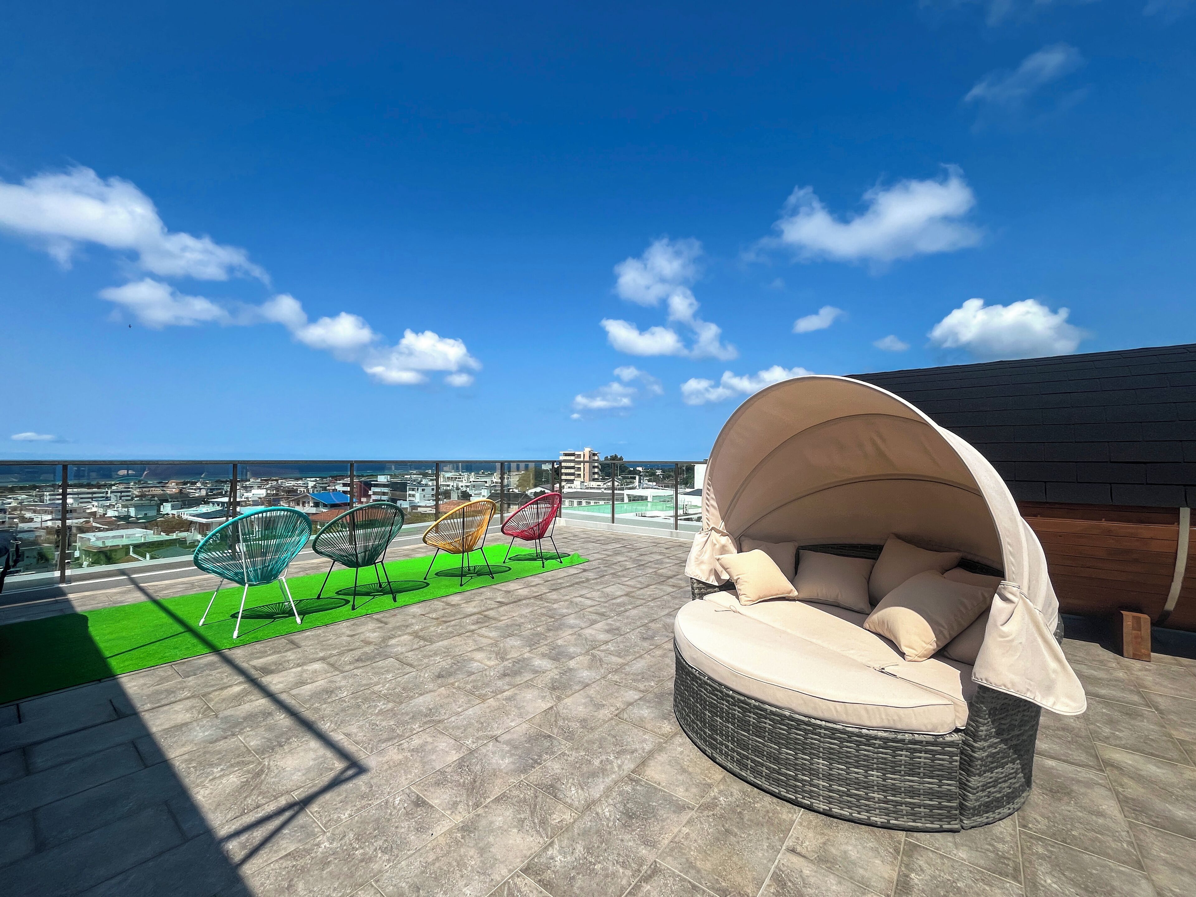 A rooftop terrace with a wicker sofa and colorful chairs, overlooking a cityscape and the sea.