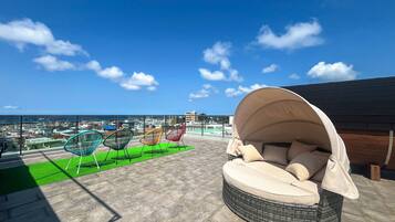 A rooftop terrace with a wicker sofa and colorful chairs, overlooking a cityscape and the sea.