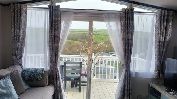 A room with a sofa, a TV, and a view of a deck and greenery outside. - Luxury caravan, Newquay Bay. Amazing views. (Newquay )