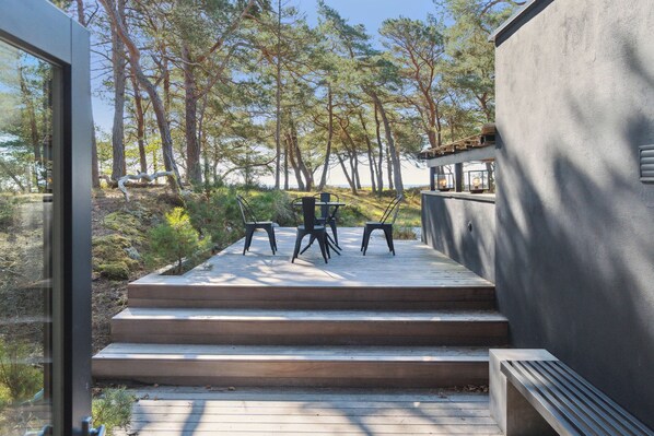 Un área de estar exterior moderna con una mesa y sillas, rodeada por una terraza de madera y vistas a árboles.