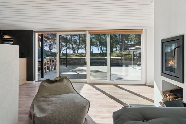 Un salon moderne doté d’une grande porte-fenêtre coulissante donnant sur une terrasse extérieure, d’une cheminée et d’un coin salon avec un fauteuil poire et un canapé.
