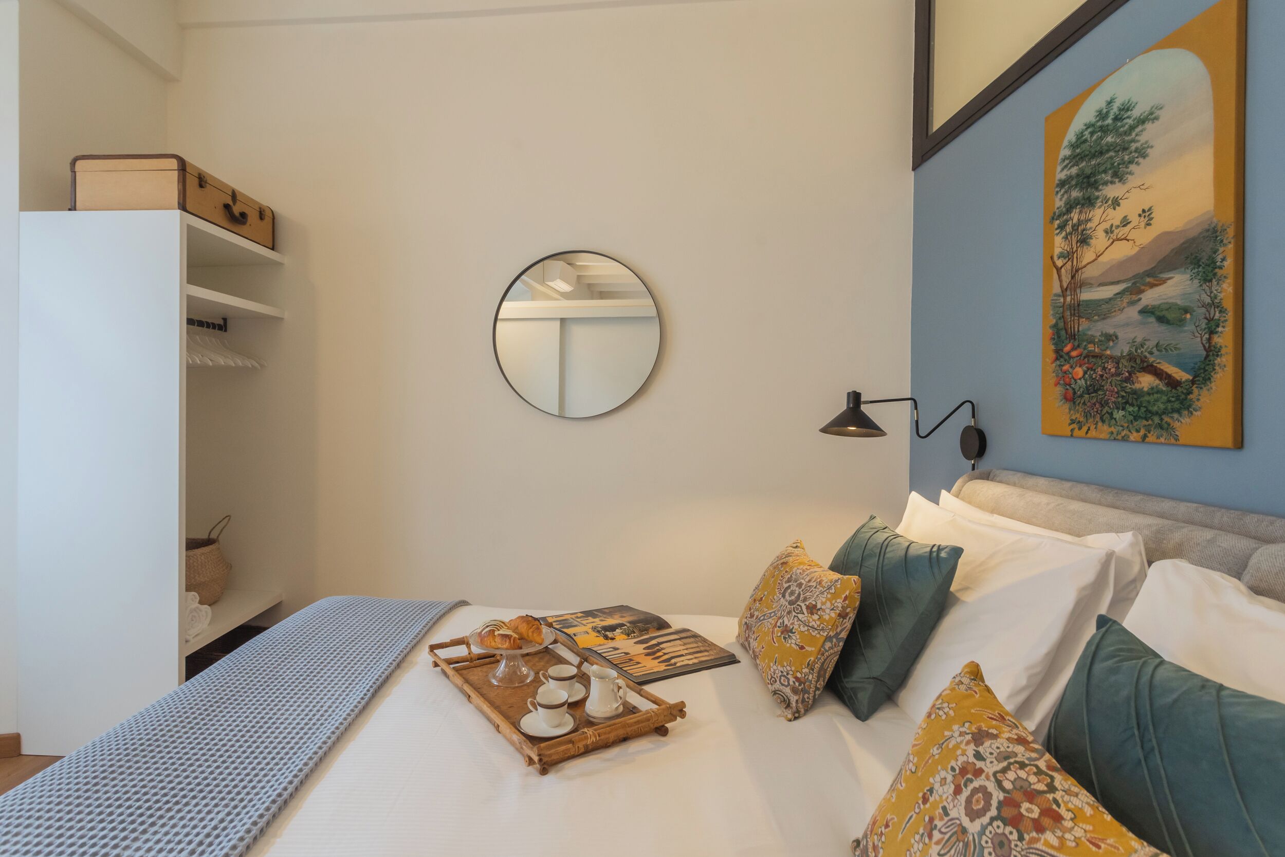 A neatly arranged bedroom with a bed, pillows, a tray with breakfast items, a mirror, and a painting on the wall.