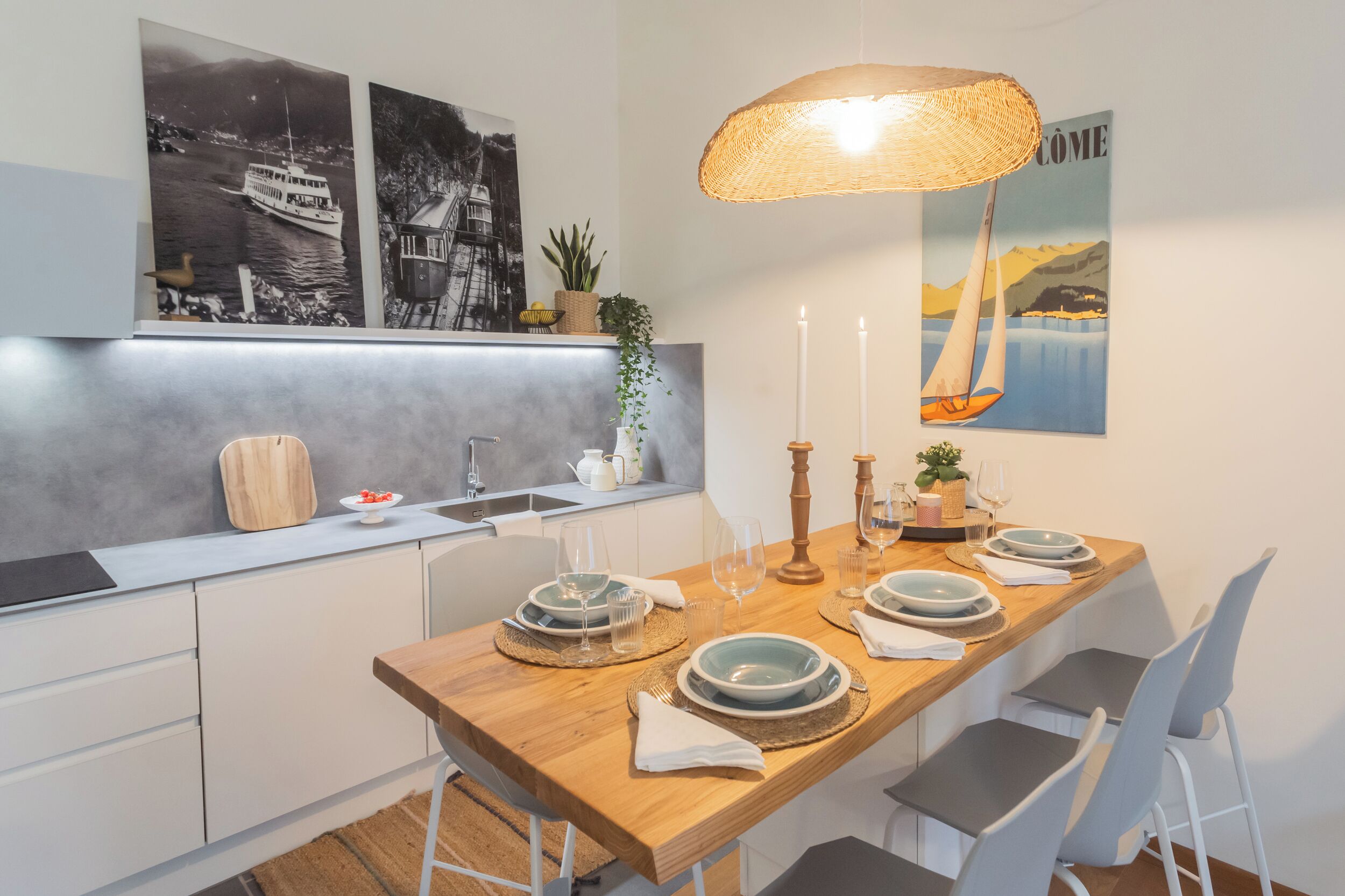 A modern kitchen with a wooden dining table set for four, a hanging wicker lamp, and artwork depicting boats and mountains.