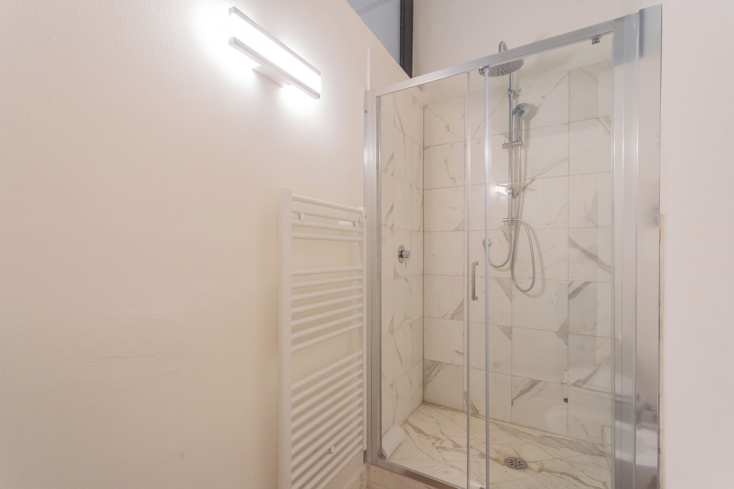 Ducha con cerramiento de vidrio, interior con azulejos de mármol, radiador blanco y lámpara en el techo.