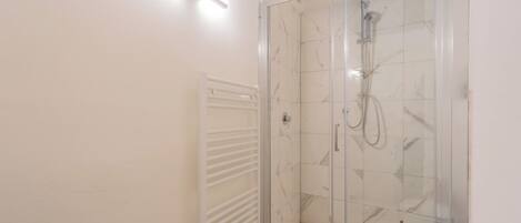 Ducha con cerramiento de vidrio, interior con azulejos de mármol, radiador blanco y lámpara en el techo.