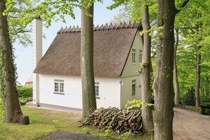 Une maison traditionnelle avec un toit de chaume, entourée d’arbres et d’une pile de bûches.