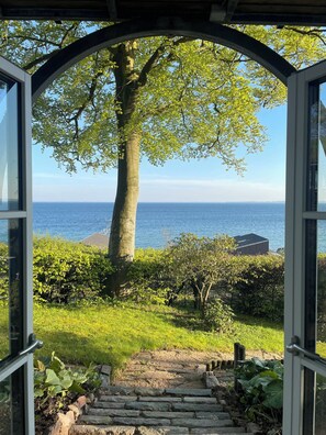 Une vue à travers une porte ouverte, donnant sur un jardin avec un grand arbre et une étendue d’eau en arrière-plan.