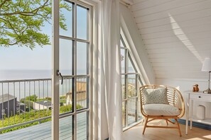 A balcony with a view of the sea, a wicker chair with a cushion, and a white desk with a lamp.