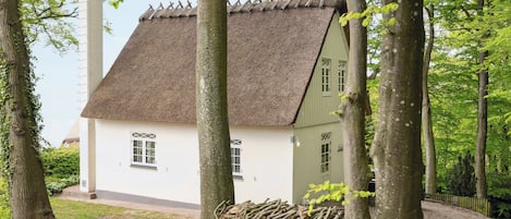 A traditional house with a thatched roof, surrounded by trees and a pile of logs.