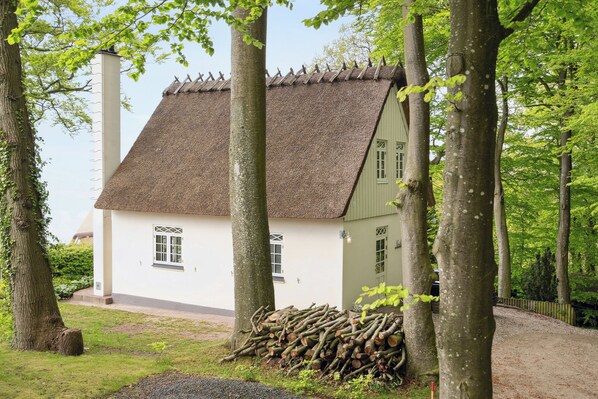 Une maison traditionnelle avec un toit de chaume, entourée d’arbres et d’une pile de bûches.