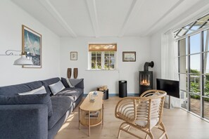 Un salon comprenant un canapé, une table, un fauteuil en osier, une cheminée et une grande fenêtre.